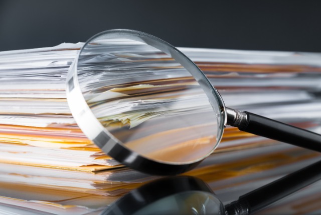 書類の山に置かれた虫眼鏡。雇用契約や在留資格など、特定技能外国人の書類確認をイメージした写真。