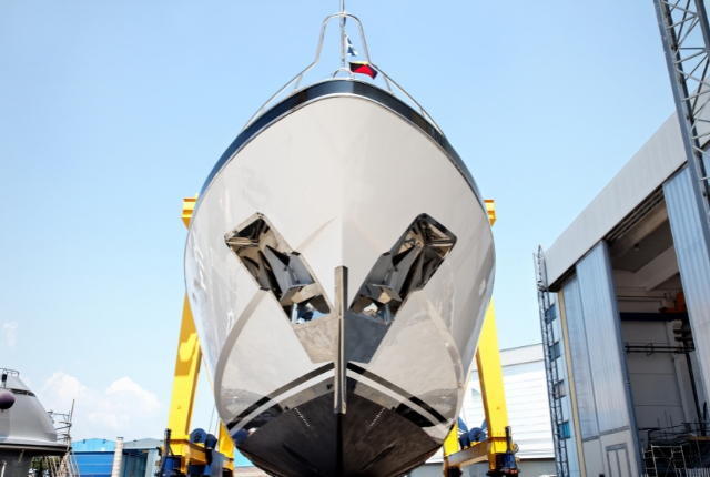 造船所で建造中の大型船の船首部分。艤装作業中の艦首がクレーンで支えられ、青空の下で整備されている様子。