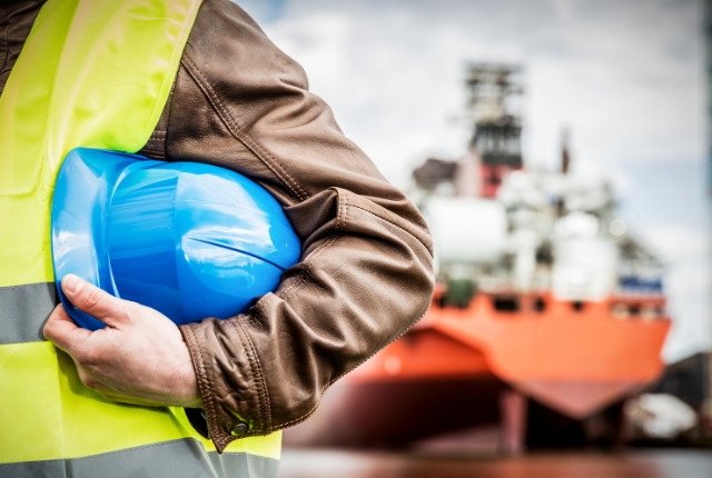 造船所で青いヘルメットを持つ作業員。安全ベストを着用し、背後には建造中の大型船が見える現場の様子。