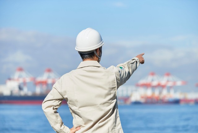 港で作業指示を出すヘルメット姿の作業員。造船所や海上現場での安全管理とチーム運営をイメージ。
