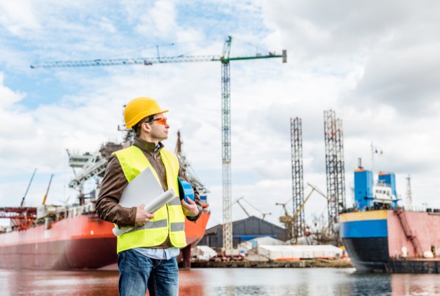 造船所で作業計画を確認する技術者。ヘルメットと図面を手に、安全ベストを着用して船の前に立つ姿