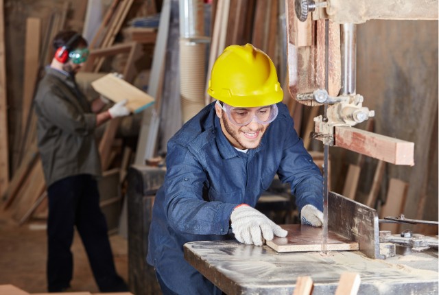木材加工作業 木工作業場で安全帽と保護メガネを着けた作業員が電動のこぎりで木材を加工する様子。製材や木工の現場写真。