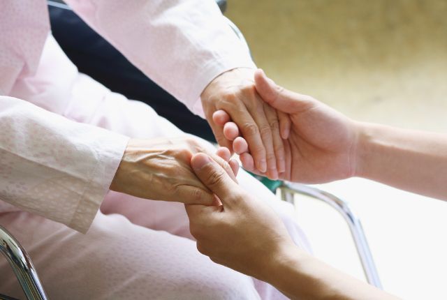 介護業界での事例紹介 車椅子の高齢者の手を介護士が優しく握る様子。思いやりや介護の温かい支援を表すイメージ。
