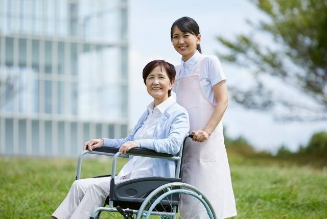 介護職員が屋外で車椅子の高齢者を支援する様子。明るい笑顔で寄り添い、安心と信頼を感じさせる介護シーン。