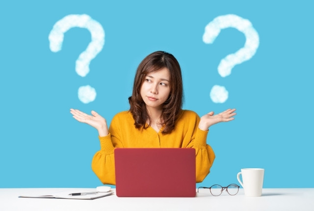 ートパソコンの前で悩む女性が両手を広げ、背景に大きなはてなマークが描かれたイメージ。制度の比較、情報収集に迷う様子を表現している。