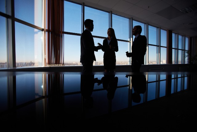 英語での商談 オフィスの大きな窓際で3人のビジネスパーソンが会話する。国際ビジネスや商談の打ち合わせを表すイメージ。