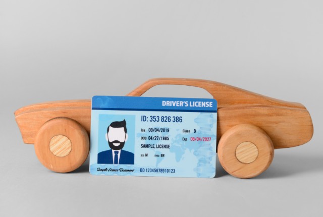木製の車の前に置かれた運転免許証の見本。自動車運送業やドライバー採用、免許取得を象徴するイメージ。