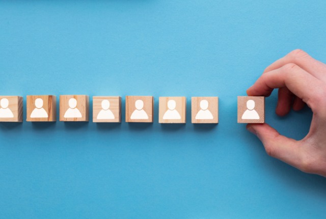 人事プロセス 青い背景の上に並べられた木製キューブに人のアイコンが描かれ、手が一つのブロックを選んでいる採用や人材選考のイメージ。