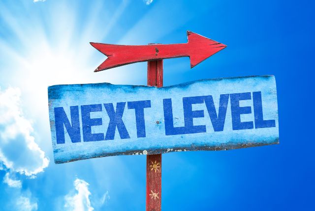 青空の下に立つ「NEXT LEVEL」と書かれた矢印看板。スキルアップや次の段階への成長を象徴。