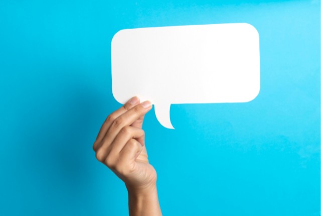 コミュニケーション 青い背景に、手が白い吹き出し型のボードを持ち上げている。英会話やコミュニケーションの象徴イメージ。