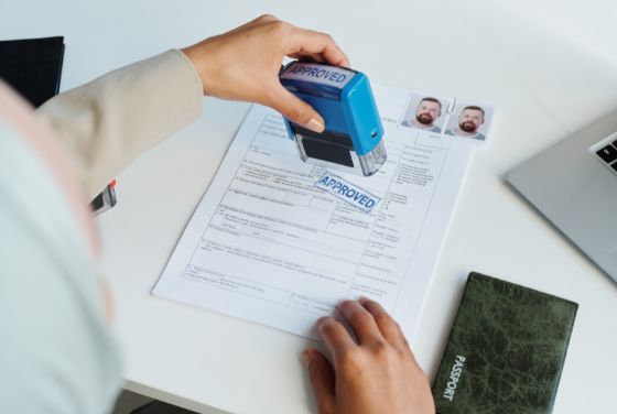 ビザ申請書に「APPROVED」スタンプを押す担当者の手元。パスポートと証明写真が並ぶ審査・承認手続きの場面。