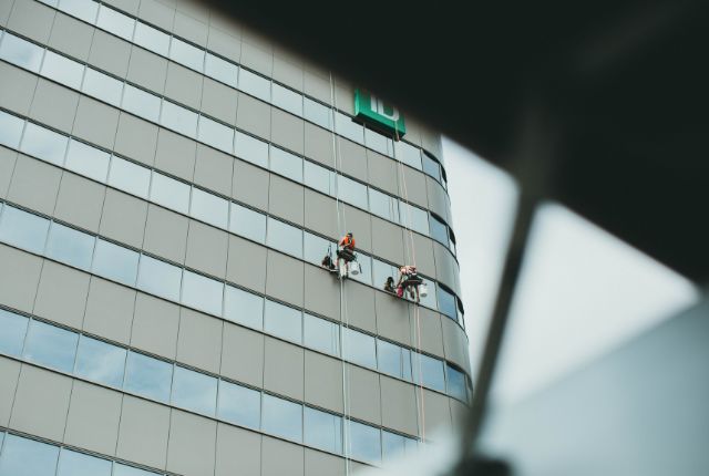 高層ビルの外壁でロープを使い窓清掃を行う作業員。ビルクリーニングや高所作業の安全管理を示す写真。