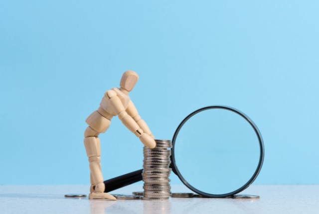 積み上げられた硬貨に木製の人形が寄りかかり、隣に虫眼鏡が置かれている。助成金や補助金、資金調査を表すイメージ。