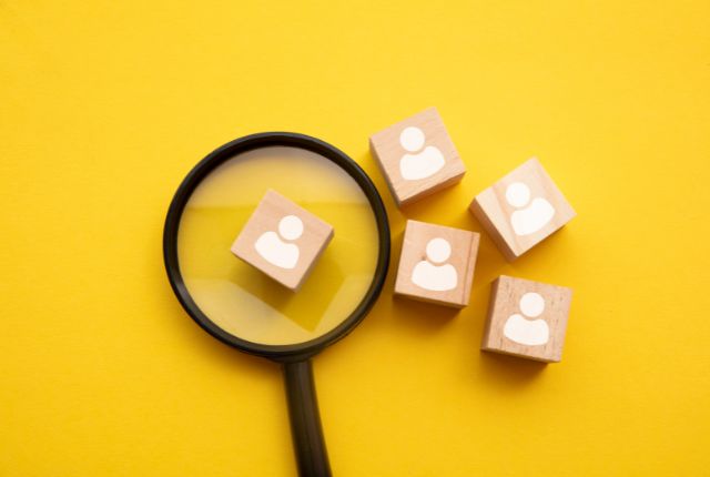 黄色い背景に置かれた虫眼鏡と人型アイコン入りの木製ブロック。採用や人材選考、外国人雇用のイメージ。