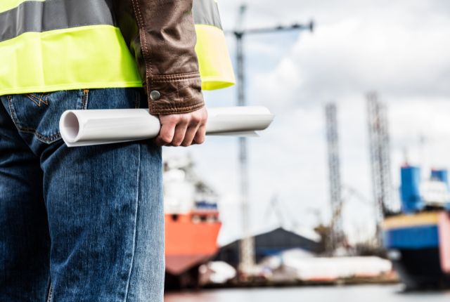 造船現場で作業員が図面を持ち、建造中の船舶を確認している様子。特定技能2号の業務イメージ。