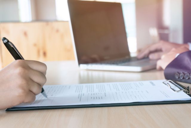 書類に記入する手元とノートパソコンが並ぶ場面を写した画像。
