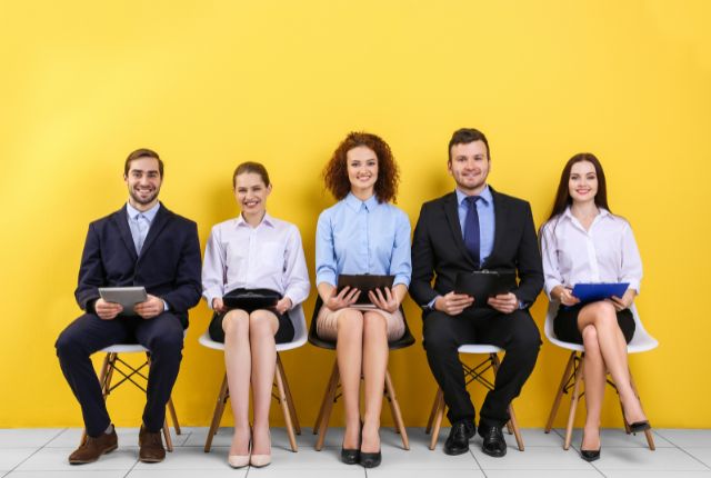 黄色背景の前でスーツ姿の応募者が並んで座る写真。採用面接や人材募集のイメージを表す。