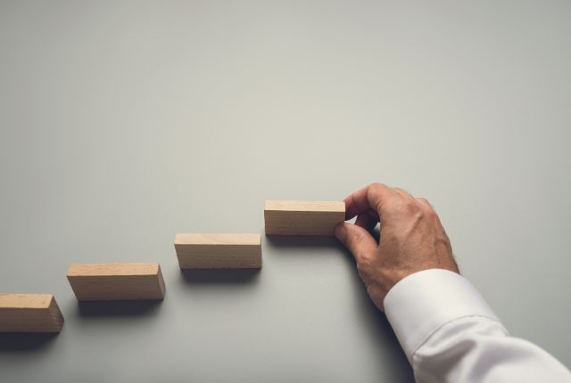 木製ブロックを階段状に積み上げる手。キャリアアップや成長、スキル向上のイメージ。