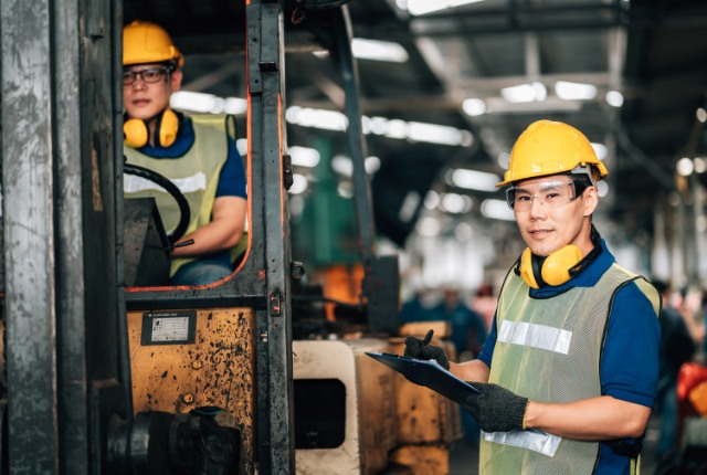 企業の事例紹介 工場内でヘルメットと安全ベストを着用した外国人作業員がフォークリフトを操作し、指示書を確認している様子。