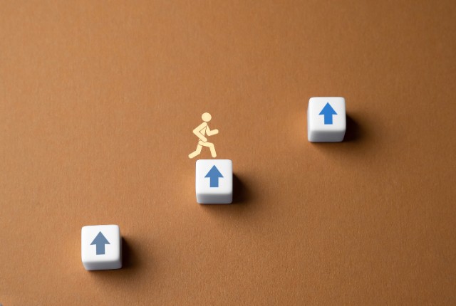上向き矢印のブロックを一段ずつ上がる人のシルエット。特定技能2号への移行やキャリアアップの象徴イメージ。