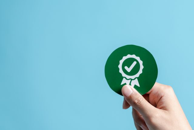 コンプライアンス 青い背景の前で、認定や合格を示すチェックマーク入りの緑色バッジを手に持つ様子。信頼性や許可取得を表現。