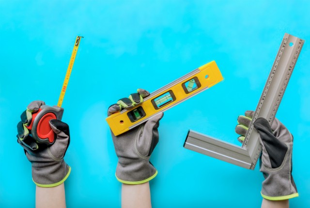 作業用手袋を着用した3本の手が、建設用のメジャー、水平器、L字定規を持っている。建設・技能実習・特定技能関連の記事に適したイメージ。