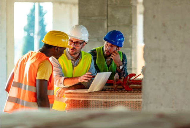 建設現場でヘルメットと安全ベストを着用した作業員が打ち合わせをしている様子。タブレットを使い作業工程を確認する特定技能関連の場面。
