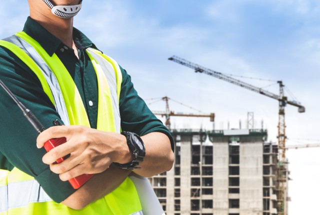建設現場を背景に安全ベストを着用し、無線機を持つ監督者の写真。工程管理や特定技能2号関連の記事に適した画像。