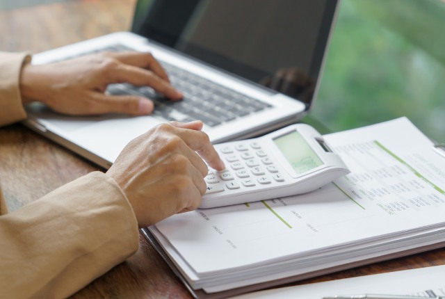 ノートパソコンで資料を確認しながら電卓を使い、技能実習生受入費用や送り出し機関の管理費を計算する人の手元