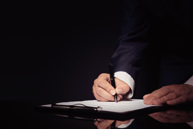 MWO申請書類への署名 暗い背景で書類に署名するスーツ姿の人物の手元を写したビジネスシーンの画像。