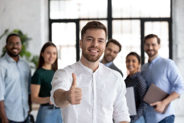 外国人従業員 職場で複数のスタッフが並び、前方の男性が親指を立てて肯定のサインを示す様子。外国人支援体制の成功イメージ。