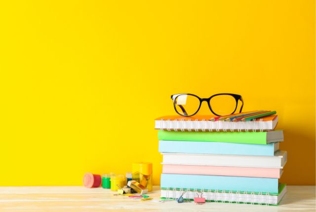 黄色背景の前にノートを重ねた文房具の置き写真。上にメガネと色鉛筆があり勉強や学習を連想させるイメージ