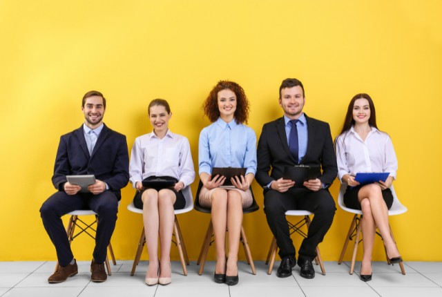 明るい黄色の壁の前で、スーツ姿の男女5人が面接を待つ様子。外国人採用・人材雇用のイメージ写真。