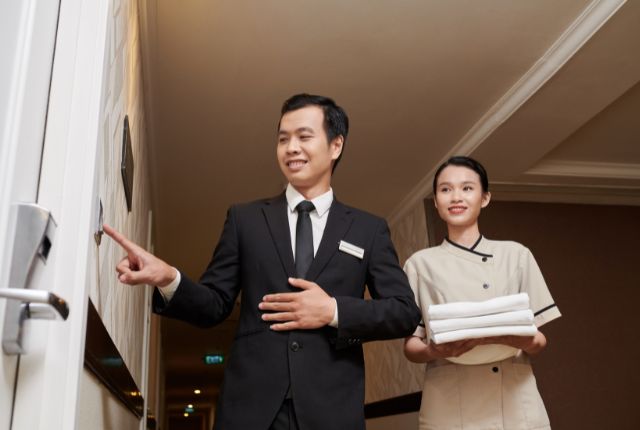 ホテル廊下でマネージャーと清掃スタッフがタオルを持って同行している研修場面