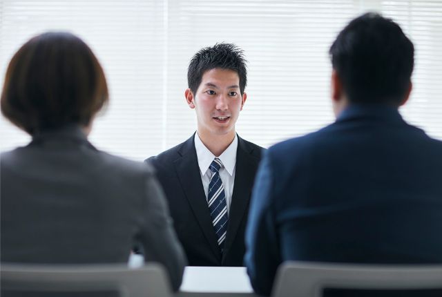新卒外国人採用の面接シーン。スーツ姿の学生が企業担当者と向き合い、留学生採用の流れを説明する場面。