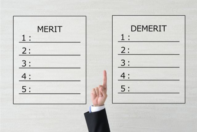 スーツ姿の手が指を立て、メリットとデメリットを比較する2つの表を指し示す。外国人雇用の利点と課題の説明イメージ。