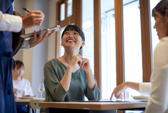レストランで注文を取る店員と笑顔で会話する女性客。外食業の接客スキルやサービス品質のイメージ。