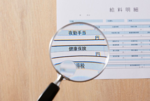 拡大鏡で給与明細書の「健康保険」「夜勤手当」などを確認する様子。外国人雇用時の社会保険・給与管理のイメージ。