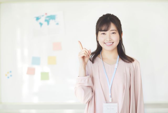 ホワイトボード前で指を立てて説明する女性スタッフの写真。採用ポイントや案内を示すイメージ。