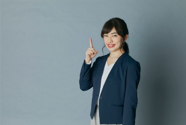 スーツ姿の女性が指を立ててポイントを示すポーズ。ビジネスでの説明やアドバイスをイメージした写真。
