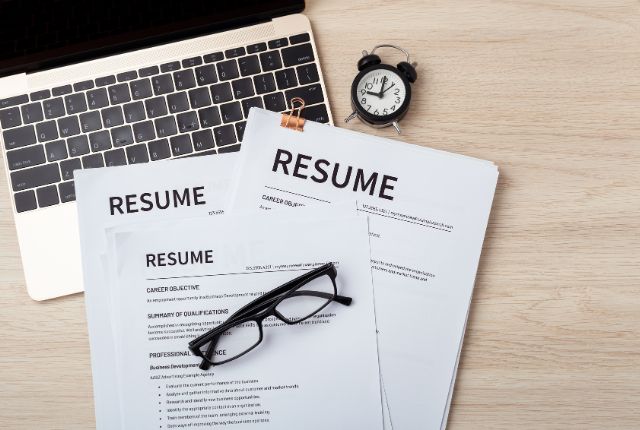 ノートパソコンと複数の履歴書が並ぶ机の上の様子。留学生採用や新卒外国人採用に必要な書類準備を示すイメージ。