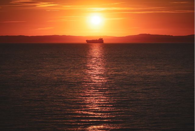 夕日が海に沈む中、水平線上を航行する船のシルエット。