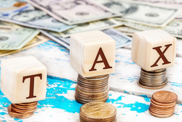 積み重ねたコインと紙幣の上に「TAX」と書かれた木製ブロックが並ぶ、税金や消費税をイメージした写真