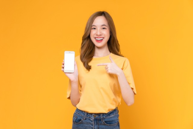 スマホ画面を指差して紹介する女性。明るい黄色背景でアプリ利用をイメージ。