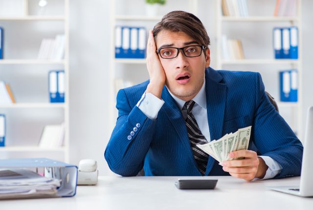 オフィスで悩んだ表情の男性が紙幣を持つ写真。給与や支出の不安、賃金トラブルを表すイメージ。